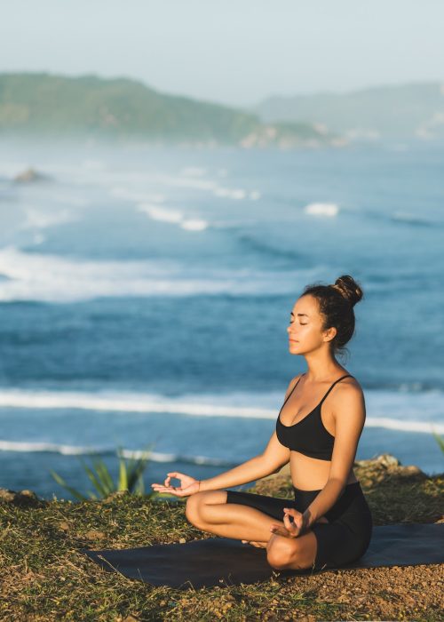 Woman practicing yoga and sit in lotus pose outdoor with amazing ocean view. Health and well-being
